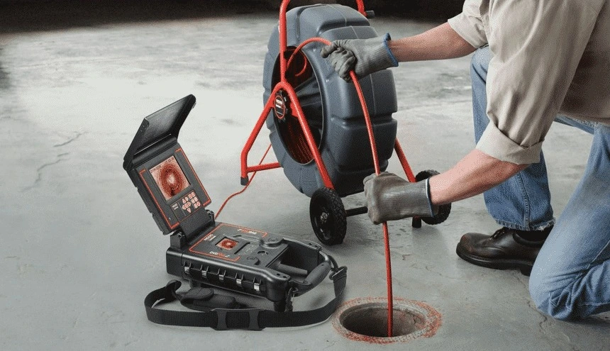 A technician feeds a cable into a floor drain for a sewer camera inspection, while monitoring a portable video screen.